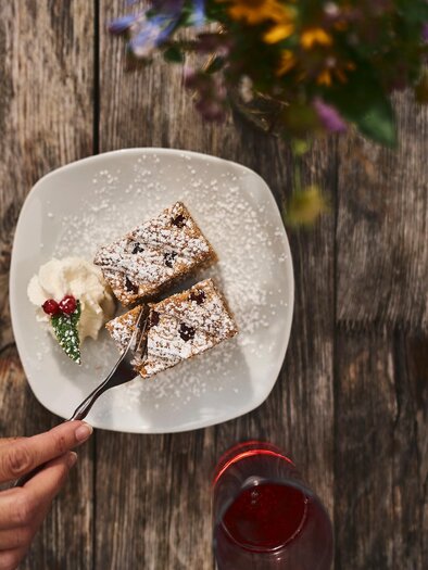Ein Teller mit zwei Stück Kuchen, bestäubt mit Puderzucker, liegt auf einem Holztisch. Daneben befindet sich eine Portion Sahne und ein Glas mit rohem Getränk. | © TV Gesäuse