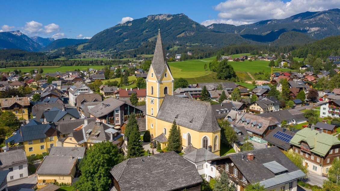 Vogelperspektive auf die gelbe Pfarrkirche mit schönem Blick auf den Ortskern von Bad Mitterndorf. Der strahlend blaue Himmel unterstreicht die sommerliche Stimmung an diesem schönen Tag. | © Karl Grieshofer