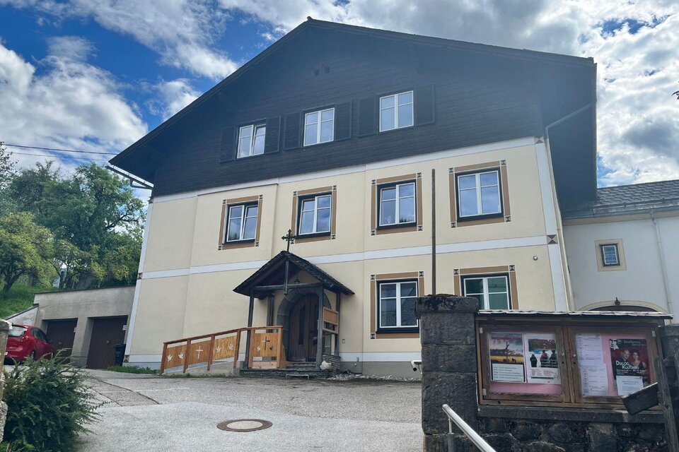 Das gelbe Pfarrhaus von Bad Mitterndorf mit dunklem Holz im obersten Geschoss und einem Kreuz über dem Eingang. Davor befindet sich ein großer asphaltierter Platz, umgeben von Bäumen. Darüber strahlt ein blauer, leicht bewölkter Himmel. | © Annika Kamp