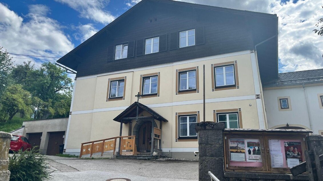 Das gelbe Pfarrhaus von Bad Mitterndorf mit dunklem Holz im obersten Geschoss und einem Kreuz über dem Eingang. Davor befindet sich ein großer asphaltierter Platz, umgeben von Bäumen. Darüber strahlt ein blauer, leicht bewölkter Himmel. | © Annika Kamp