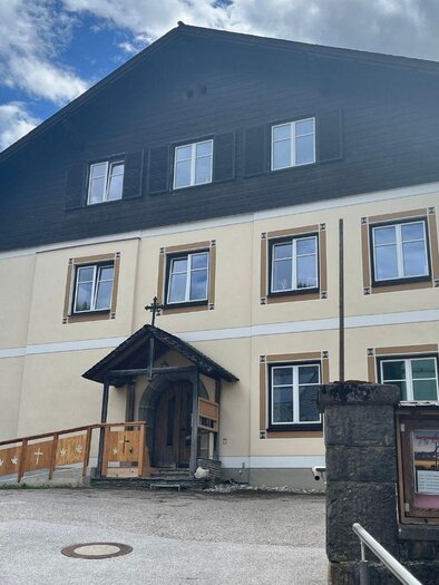 The yellow rectory of Bad Mitterndorf with dark wood on the top floor and a cross above the entrance. In front of it is a large asphalted square, surrounded by trees. Above it shines a blue, slightly cloudy sky. | © Annika Kamp