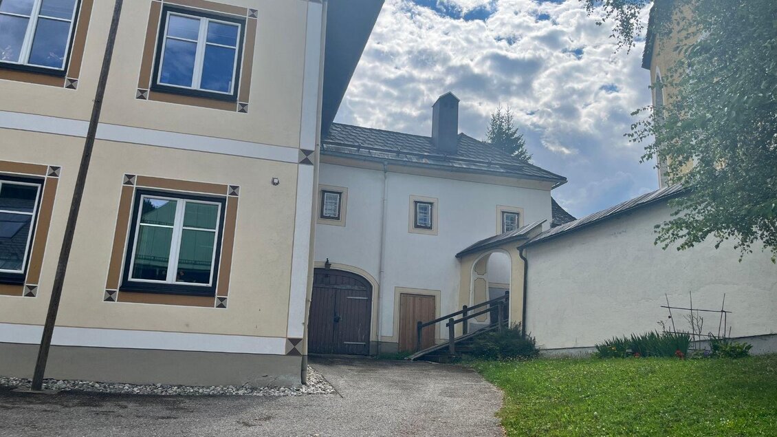 Neben dem Pfarrhaus führt eine Treppe zur Kirche, die teilweise durch einen Baum sichtbar ist. Neben dem asphaltierten Platz befindet sich eine kleine grüne Fläche. | © Annika Kamp