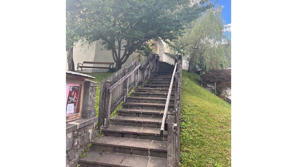 Eine Steintreppe führt zur Kirche, rechts davon erstreckt sich eine Wiese, während links oben eine Bank zwischen zwei Bäumen steht. Vor der Treppe befindet sich ein Schaukasten mit aktuellen Informationen. | © Annika Kamp