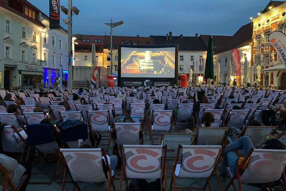 Ein Freiluftkino mit vielen Liegestühlen, die vor einer großen Leinwand aufgestellt sind. Die Szene spielt abends in einer Stadt mit historischem Ambiente. | © Silent Cinema