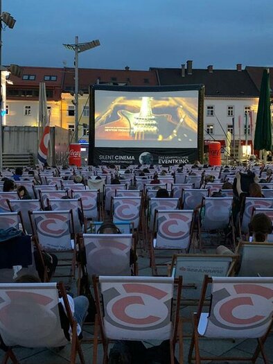Ein Freiluftkino mit vielen Liegestühlen und einem großen Bildschirm. Die Veranstaltung findet bei Abendlicht in einer Stadtplatzatmosphäre statt. | © Bild: Silent Cinema
