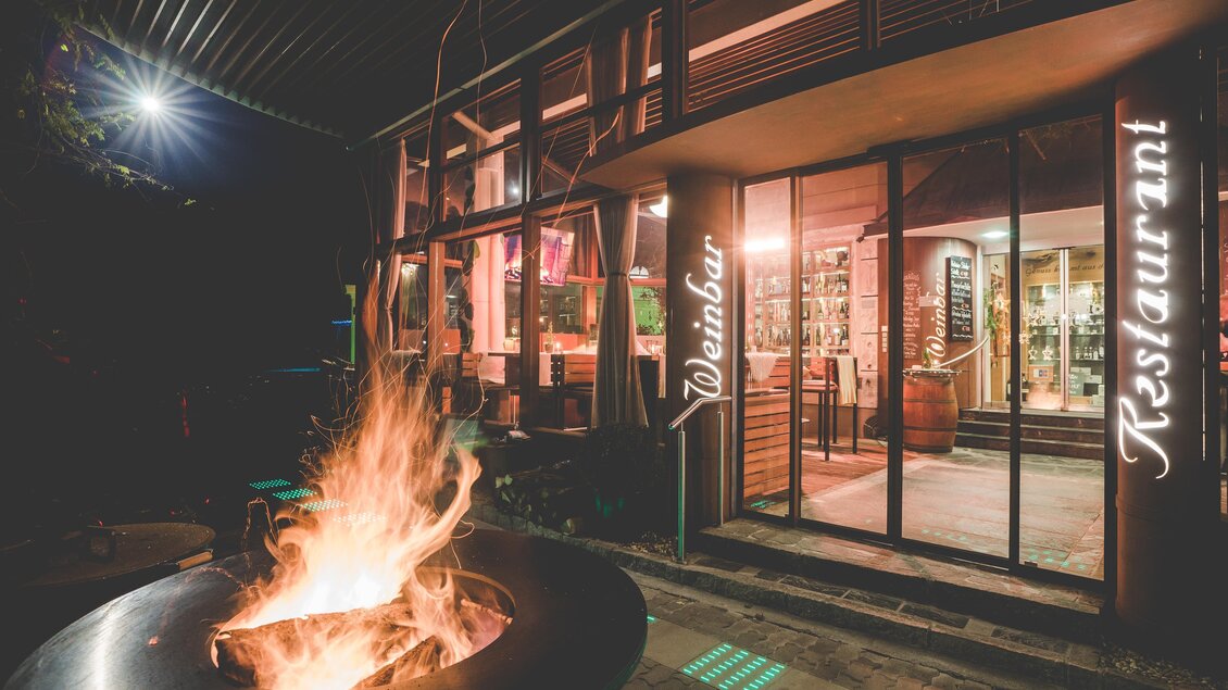 Ein einladendes Restaurant mit großen Fenstern und warmem Licht. Vor dem Gebäude brennt ein gemütliches Feuer. | © Garten Hotel Ochensberger