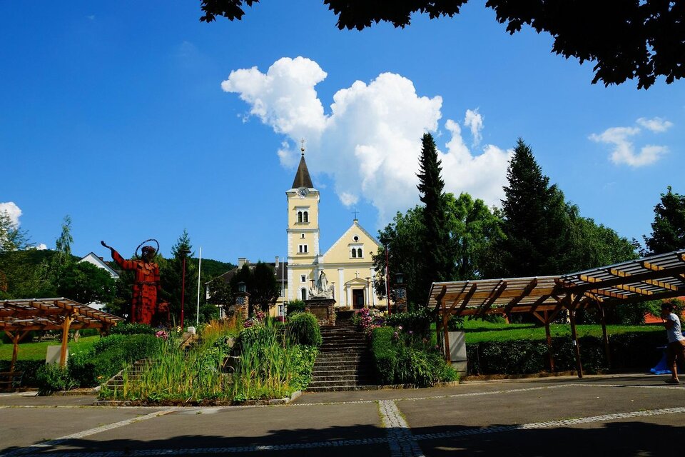 Eine malerische Kirche in einer grünen Umgebung unter einem blauen Himmel. Im Vordergrund sind Parks und kunstvolle Installationen zu sehen. | © TV Thermen- & Vulkanland
