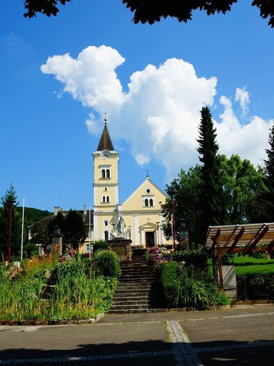 A picturesque church in a green setting under a blue sky. In the foreground, parks and artistic installations can be seen. | © TV Thermen- & Vulkanland
