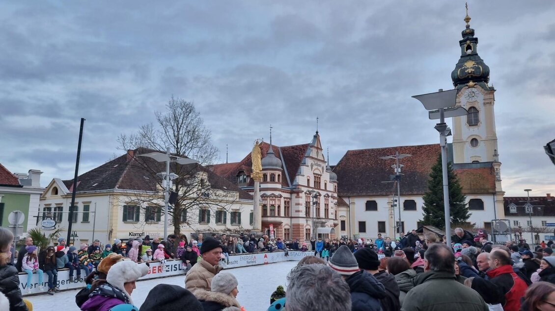 Eine Eislauffläche mit vielen Menschen in einer Stadt. Im Hintergrund sind historische Gebäude und ein Kirchturm zu sehen. | © Stadtmarketing Hartberg
