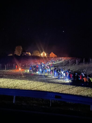 Eine große Gruppe von Menschen wandert bei Nacht entlang eines Feldes. Im Hintergrund sind beleuchtete Häuser zu sehen. | © Hannes Kofler