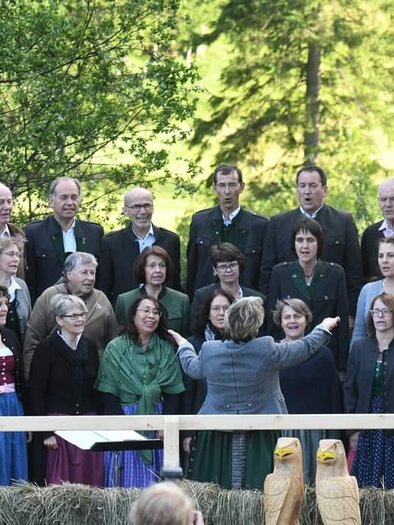 A group of singers stands on a stage amidst trees. They are wearing traditional clothing and are being conducted by a conductor. | © TVB Murau