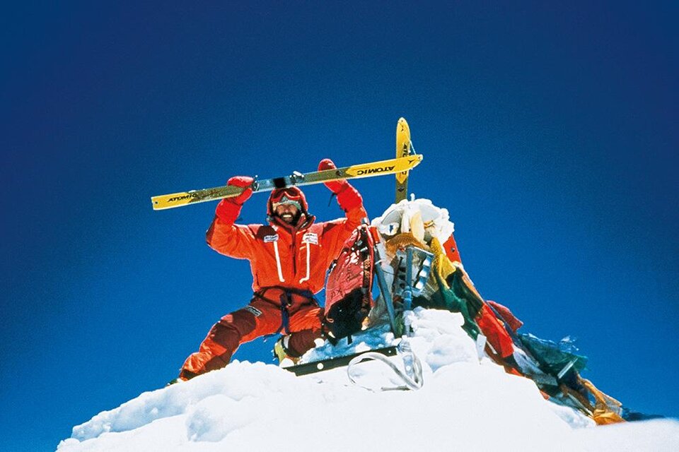 Ein Mann steht auf einem schneebedeckten Gipfel und hält ein Paar Skier über seinen Kopf. Der Himmel ist klar und blau, was die beeindruckende alpine Szene unterstreicht. | ©  Kammerlander