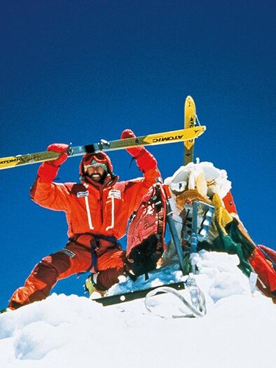 A man stands on a snow-covered summit, holding a pair of skis above his head. The sky is clear and blue, highlighting the impressive alpine scene. | ©  Kammerlander