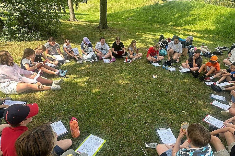 Schüler sitzen im Kreis auf einer Wiese im Skulpturenpark Premstätten und arbeiten mit Unterlagen im Freien. | © Kunst- und Naturvermittlung UMJ