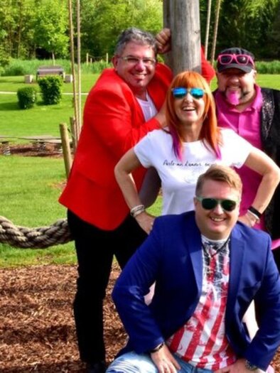 Eine Gruppe von vier fröhlichen Personen posiert in einem Park. Sie tragen bunte Kleidung und lächeln in die Kamera, während sie vor einer Spielplatzschaukel stehen. | © DaLuis