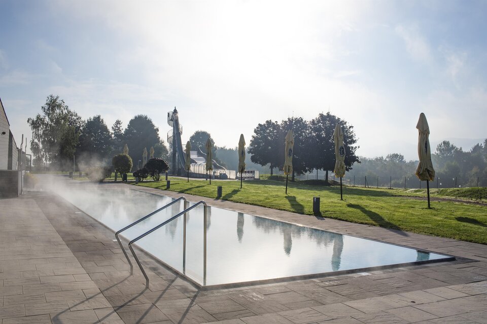 Ein ruhiger Pool mit dampfendem Wasser und einer grünen Umgebung. Sonnenschirme stehen bereit, während die Sonne langsam aufgeht. | © Anita Fössl