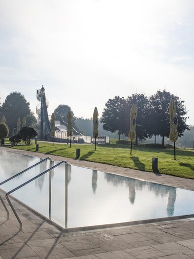 A quiet pool with steaming water and a green environment. Sun umbrellas are ready as the sun slowly rises. | © Anita Fössl