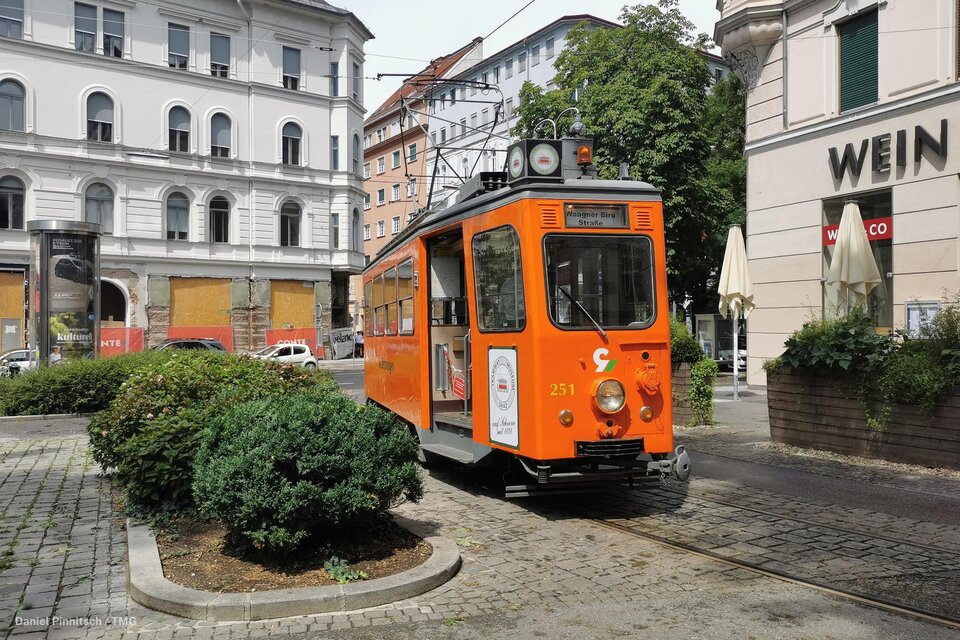 Eine orange Straßenbahn steht an einer Haltestelle in der Stadt. Im Hintergrund sind alte Gebäude und Bäume zu sehen. | © _tramway museum graz