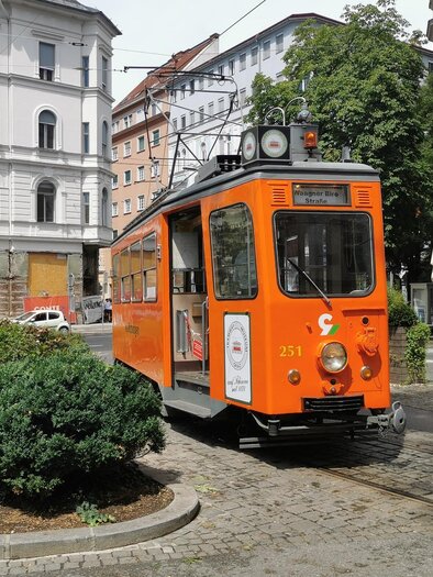 Eine orange Straßenbahn steht an einer Haltestelle in der Stadt. Im Hintergrund sind alte Gebäude und Bäume zu sehen. | © _tramway museum graz