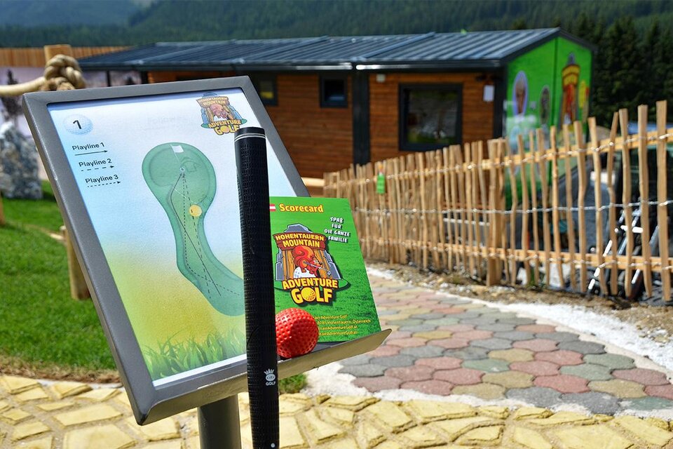 Eine Karte für Adventure Golf mit einem Schläger und einem Ball im Vordergrund. Im Hintergrund sieht man ein Holzgebäude und einen gepflasterten Weg. | © Mountain Adventure Golf