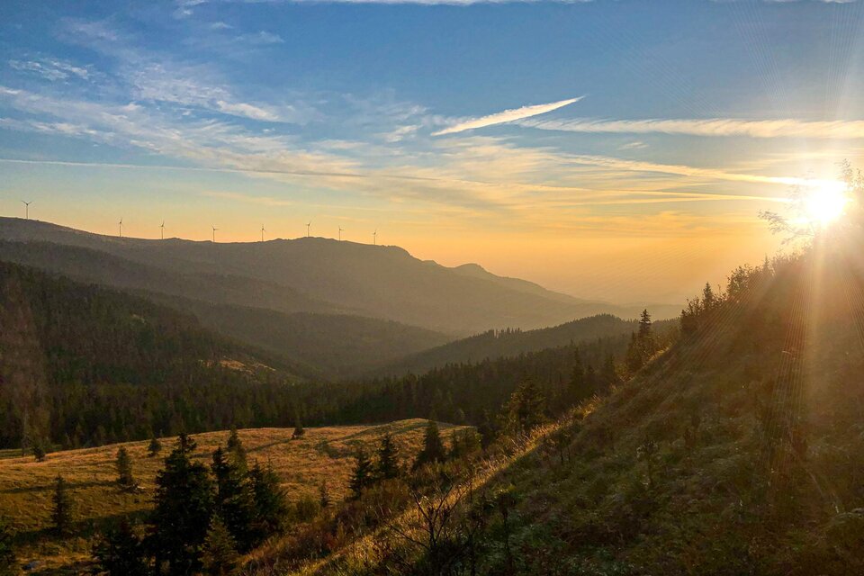 Eine malerische Berglandschaft mit Sonnenuntergang und sanften Hügeln. Im Hintergrund sind Windräder und der Himmel ist blau mit einigen Wolken. | © TV Südsteiermark_Christian Freydl