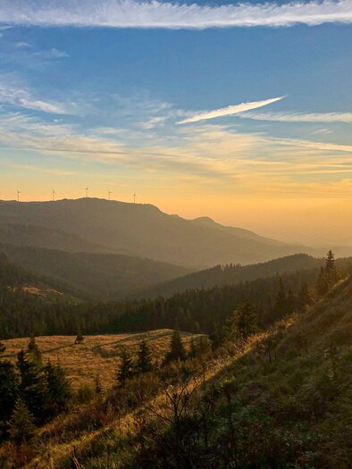 Eine malerische Berglandschaft mit Sonnenuntergang und sanften Hügeln. Im Hintergrund sind Windräder und der Himmel ist blau mit einigen Wolken. | © TV Südsteiermark_Christian Freydl