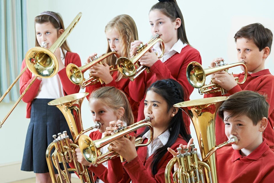 Eine Gruppe von Kindern spielt gemeinsam Trompete und Posaune. Sie tragen rote Pullover und konzentrieren sich auf ihre Musik.