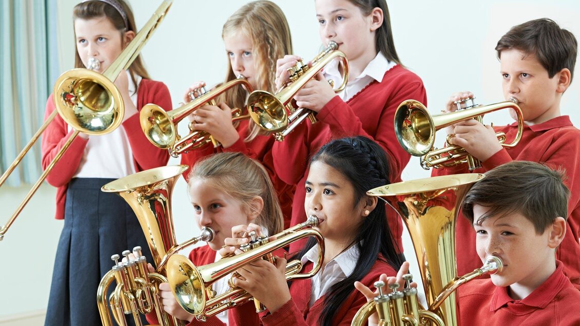 Eine Gruppe von Kindern spielt gemeinsam Trompete und Posaune. Sie tragen rote Pullover und konzentrieren sich auf ihre Musik.