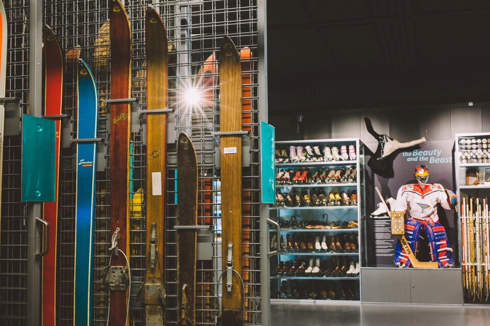 Eine Innenansicht eines Sportgeschäfts mit Holzskiern und einer Wand mit Eislaufschuhen. Im Hintergrund ist ein Plakat mit einem Sportler zu sehen. | © nicoleseiser.at
