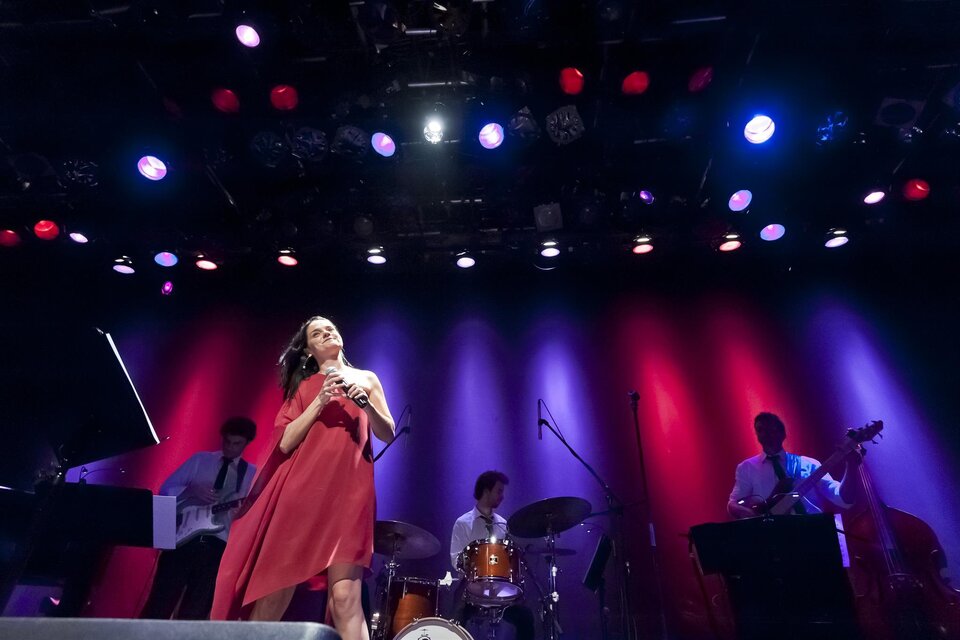Eine Sängerin auf der Bühne in einem roten Kleid, umgeben von Musikern. Bunte Lichter strahlen im Hintergrund und schaffen eine lebendige Atmosphäre. | © Werner Krug