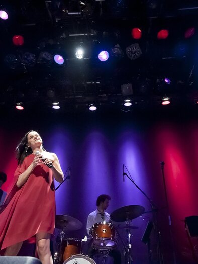 Eine Sängerin auf der Bühne in einem roten Kleid, umgeben von Musikern. Bunte Lichter strahlen im Hintergrund und schaffen eine lebendige Atmosphäre. | © Werner Krug