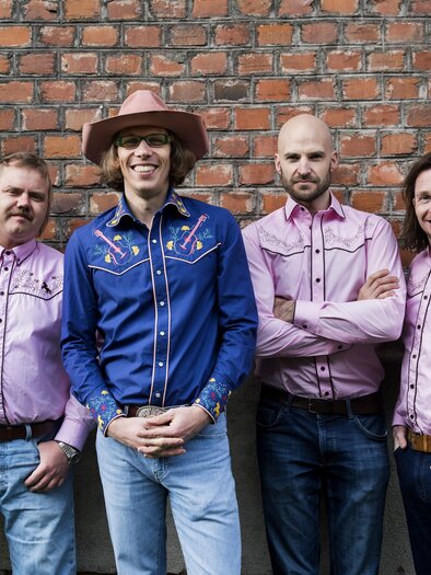 Eine Männergruppe steht vor einer Backsteinwand. Sie tragen westernähnliche Hemden und Cowboyhüte. | © Prenninger