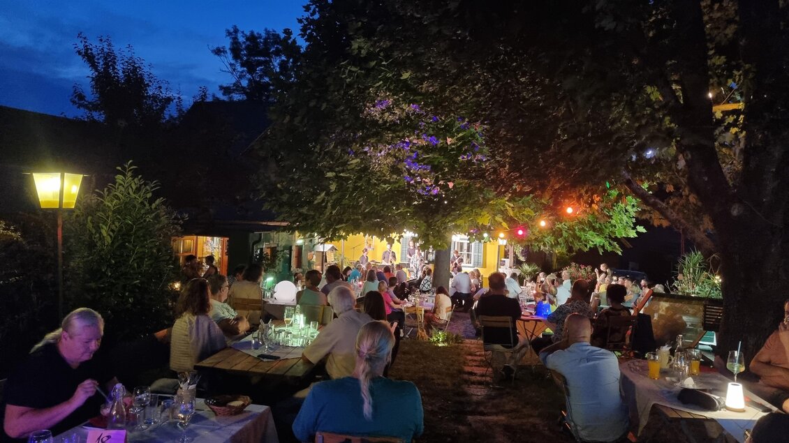 Ein belebter Außenbereich mit Tischen und Stühlen bei Nacht. Bunte Lichter strahlen von den Bäumen und schaffen eine gemütliche Atmosphäre. | © Kürzel