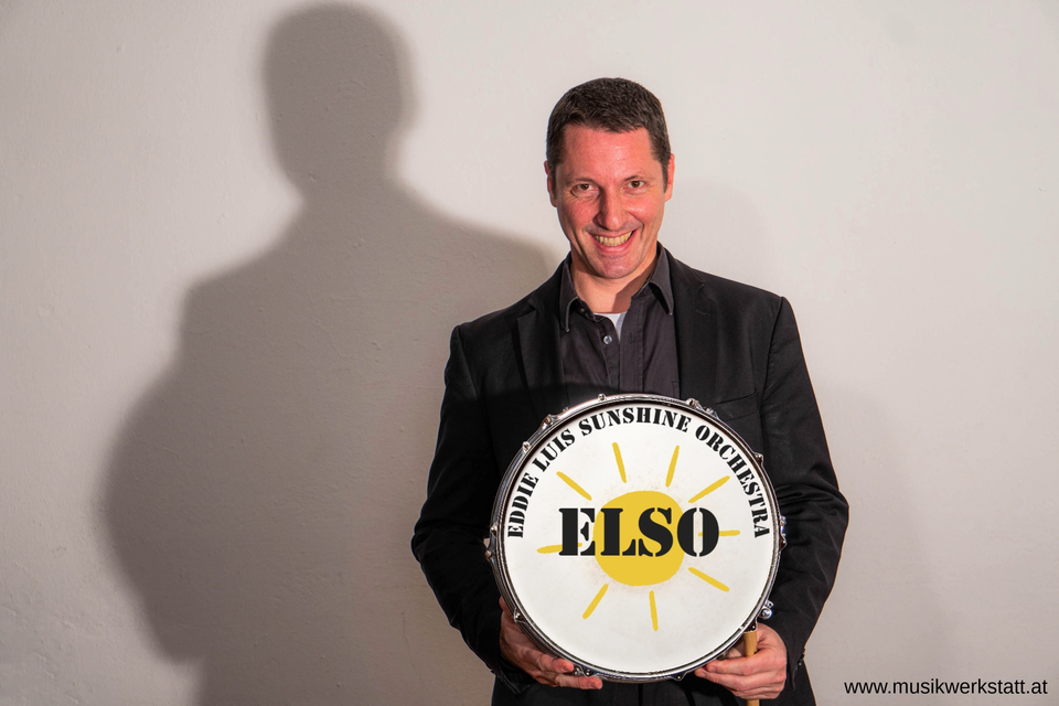 Ein lächelnder Mann hält ein Trommelschild mit der Aufschrift "ELSO". Er trägt einen schwarzen Anzug und steht vor einer hellen Wand. | © Louis