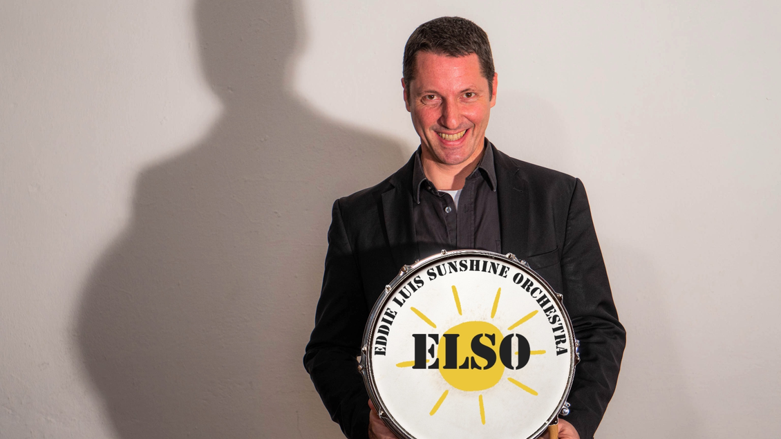 Ein lächelnder Mann hält ein Trommelschild mit der Aufschrift "ELSO". Er trägt einen schwarzen Anzug und steht vor einer hellen Wand. | © Louis
