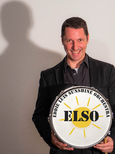 Ein lächelnder Mann hält ein Trommelschild mit der Aufschrift "ELSO". Er trägt einen schwarzen Anzug und steht vor einer hellen Wand. | © Louis