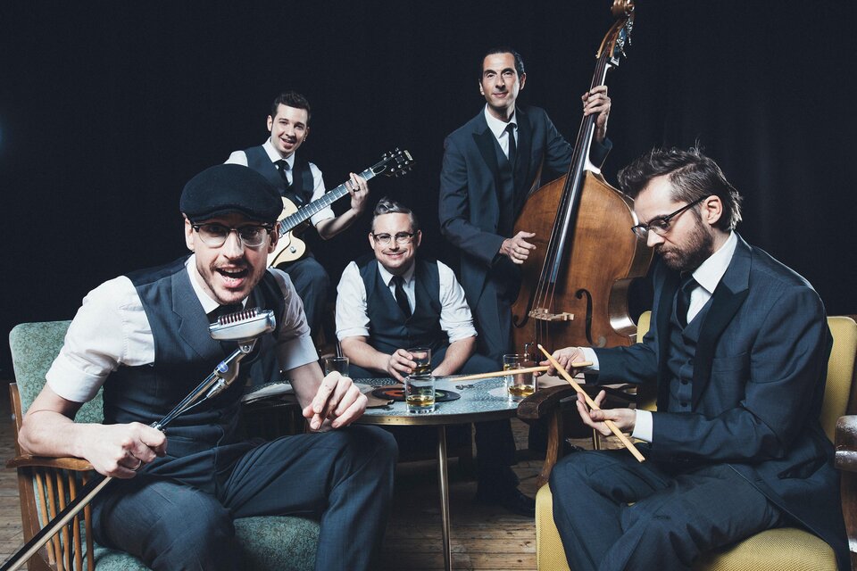 Eine Band aus fünf Männern in Anzügen spielt Musik in einem stilvollen Raum. Sie haben verschiedene Instrumente und wirken fröhlich und dynamisch. | © Marija Kanizaj