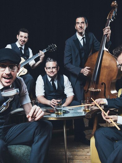 Eine Band aus fünf Männern in Anzügen spielt Musik in einem stilvollen Raum. Sie haben verschiedene Instrumente und wirken fröhlich und dynamisch. | © Marija Kanizaj