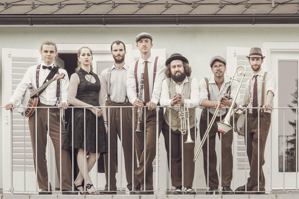 Eine Band steht auf einem Balkon und präsentiert sich in eleganter Kleidung. Sie spielen verschiedene Instrumente und lächeln in die Kamera. | © Lusija Novak