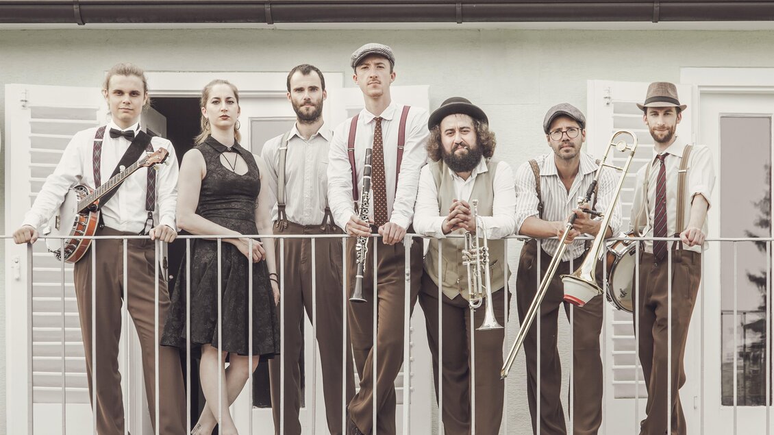 Eine Band steht auf einem Balkon und präsentiert sich in eleganter Kleidung. Sie spielen verschiedene Instrumente und lächeln in die Kamera. | © Lusija Novak