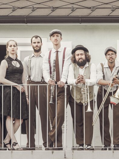 Eine Band steht auf einem Balkon und präsentiert sich in eleganter Kleidung. Sie spielen verschiedene Instrumente und lächeln in die Kamera. | © Lusija Novak