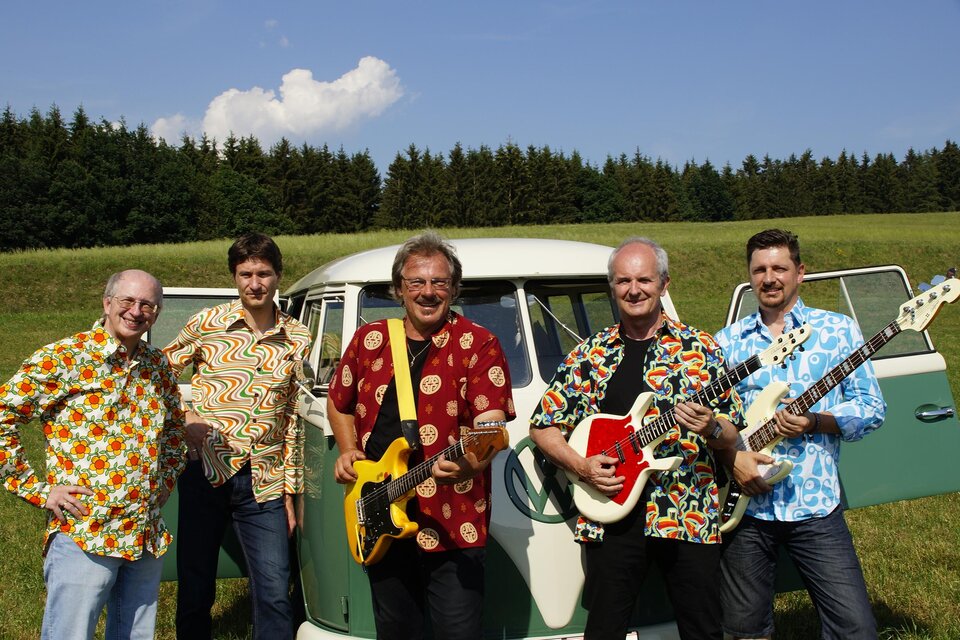 Eine Band steht vor einem Oldtimer in einem grünen Feld. Alle Mitglieder tragen bunte Hemden und halten Musikinstrumente. | © the roaring sixties company
