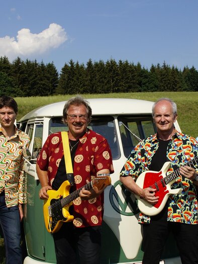 Eine Band steht vor einem Oldtimer in einem grünen Feld. Alle Mitglieder tragen bunte Hemden und halten Musikinstrumente. | © the roaring sixties company