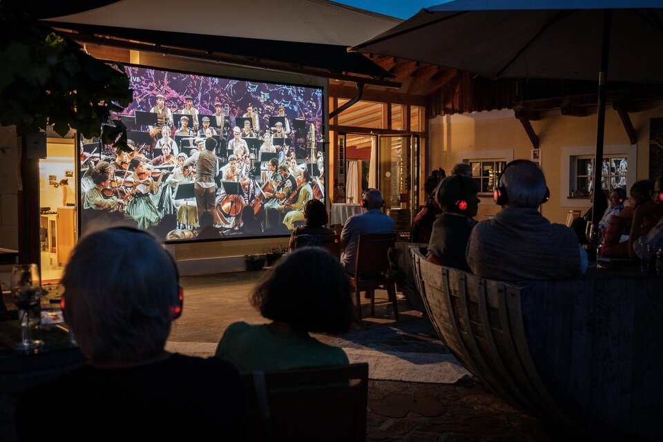 Eine Gruppe von Menschen sitzt draußen und schaut sich ein Konzert auf einer großen Leinwand an. Die Aufführung zeigt ein Orchester, das in einer stimmungsvollen Atmosphäre spielt. | © @jöbstl