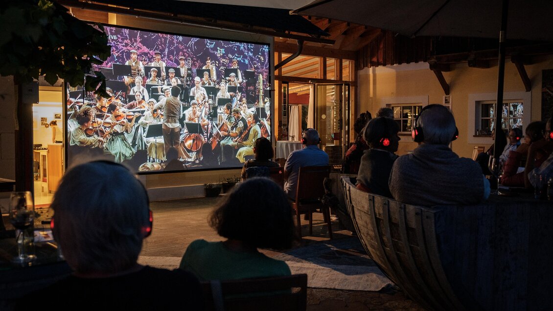 Eine Gruppe von Menschen sitzt draußen und schaut sich ein Konzert auf einer großen Leinwand an. Die Aufführung zeigt ein Orchester, das in einer stimmungsvollen Atmosphäre spielt. | © @jöbstl