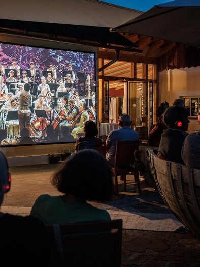 Eine Gruppe von Menschen sitzt draußen und schaut sich ein Konzert auf einer großen Leinwand an. Die Aufführung zeigt ein Orchester, das in einer stimmungsvollen Atmosphäre spielt. | © @jöbstl