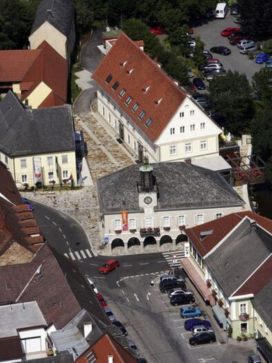 Eine Luftaufnahme einer kleinen Stadt mit verschiedenen Gebäuden und einem ruhigen Straßenverkehr. Im Mittelpunkt steht ein historisches Gebäude mit einem Uhrturm. | © Rudi Tischler