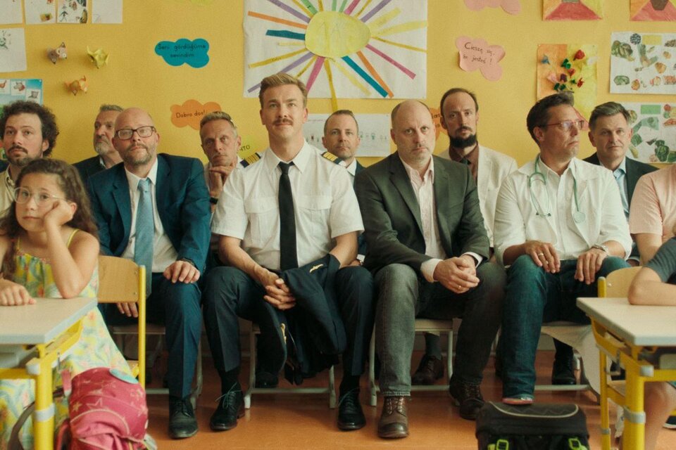 Eine Gruppe von Erwachsenen sitzt in einem Klassenzimmer, umgeben von bunten Wandbildern. Ein Kind sitzt an einem Tisch und schaut aufmerksam zu den Erwachsenen. | © Albin Wildner