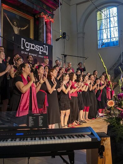 Ein Gospelchor tritt in einer Kirche auf. Die Sängerinnen und Sänger tragen schwarze und pinke Outfits und sind begeistert beim Singen. | © Peter Purgar