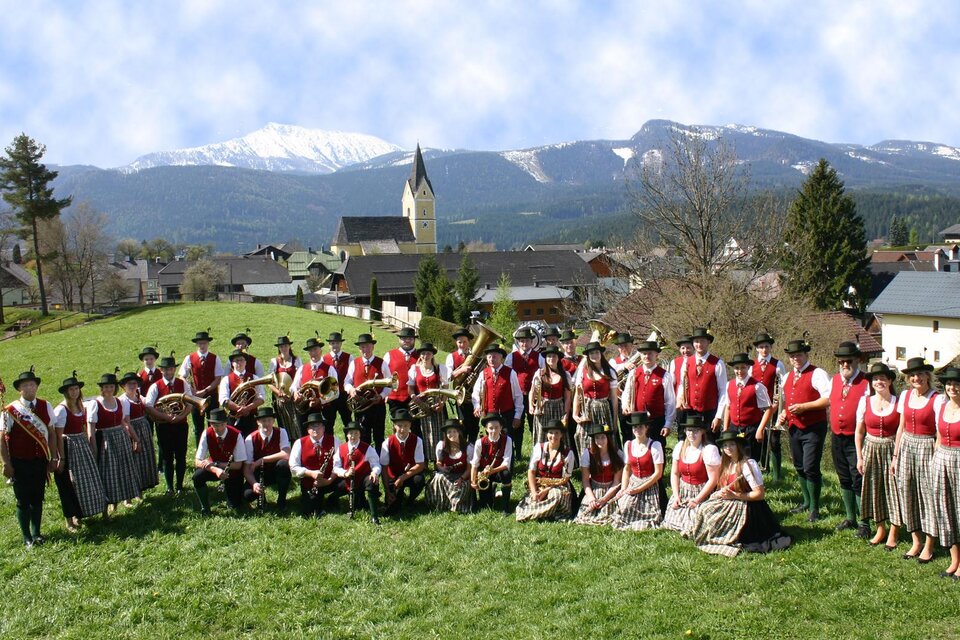 Eine große Gruppe von Menschen in traditioneller Tracht posiert auf einer Wiese. Im Hintergrund sind Berge und ein Dorf zu sehen. | © Musikkapelle Bad Mitterndorf